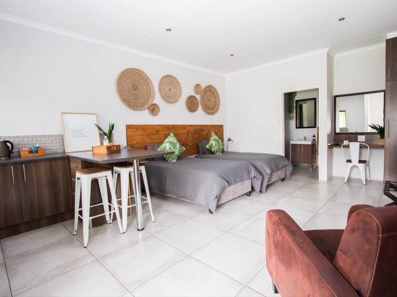 A modern, well-lit room with a kitchen area, a dining table with stools, a bed with grey bedding, and a red chair.