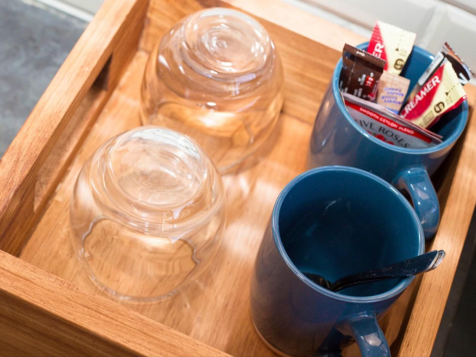A wooden tray with a clear glass pitcher, a blue mug, and a mug with a spoon.