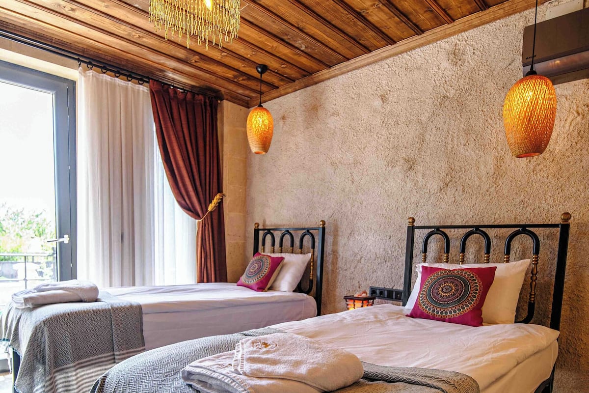 A room with two beds, wooden ceiling, and a large window with curtains.
