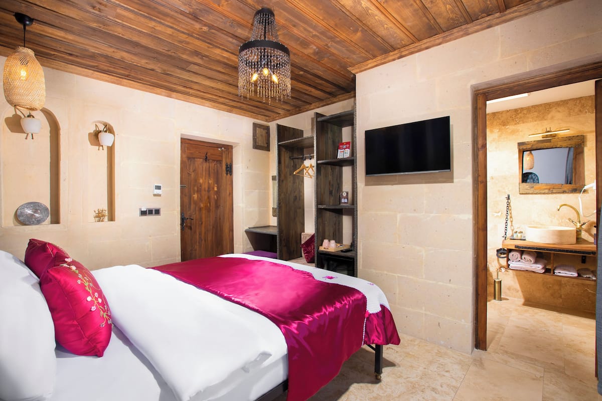 A bedroom with a bed, wooden ceiling, a chandelier, a TV, and a bathroom with a sink and mirror.