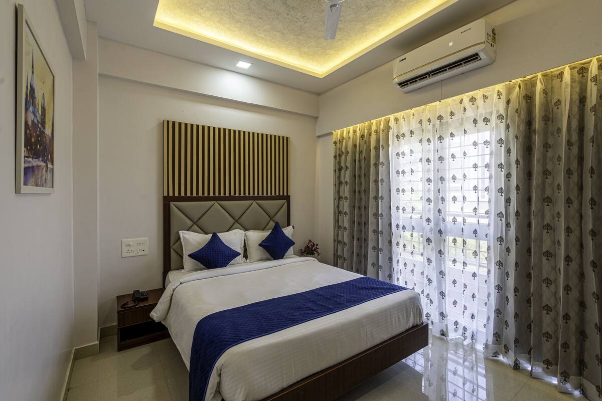 A modern hotel room with a large bed, a headboard with vertical stripes, a nightstand with a phone, and a window with patterned curtains.
