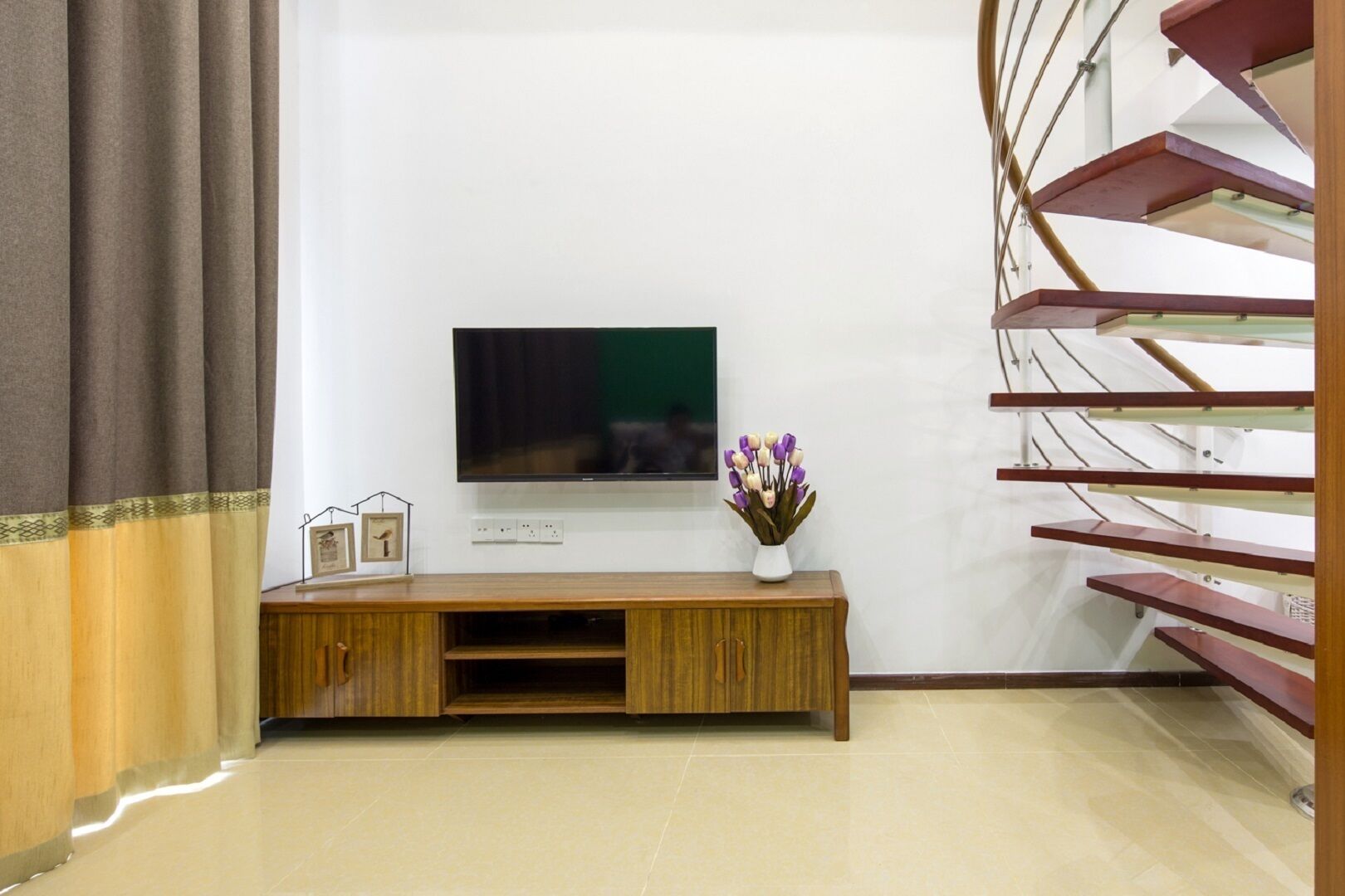 Ein modernes Wohnzimmer mit einem Flachbildfernseher auf einem Holzboden, einer Blumenvase und einer Treppe rechts.