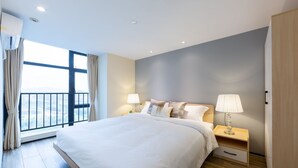A modern hotel room with a large bed, two bedside tables with lamps, a view through a large window, and a balcony.