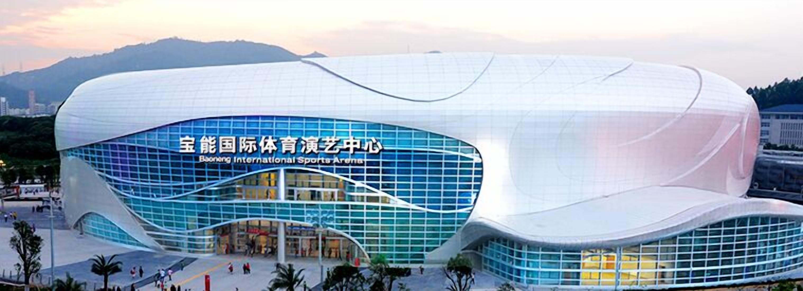 En moderne, buet bygning med en glasfacade og et skilt, der angiver, at det er Baoneng International Sports Arena.