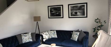 A living room with a dark blue sofa, a white coffee table, and a floor lamp. There are two framed pictures on the wall and a plant in the corner.
