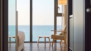 Een kamer met een bed, een stoel en een tafel, met uitzicht op de zee door grote ramen.