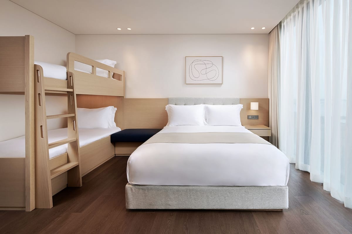 A hotel room with a bunk bed, a single bed, a bedside table with a lamp, a wall art piece, and a window with curtains.
