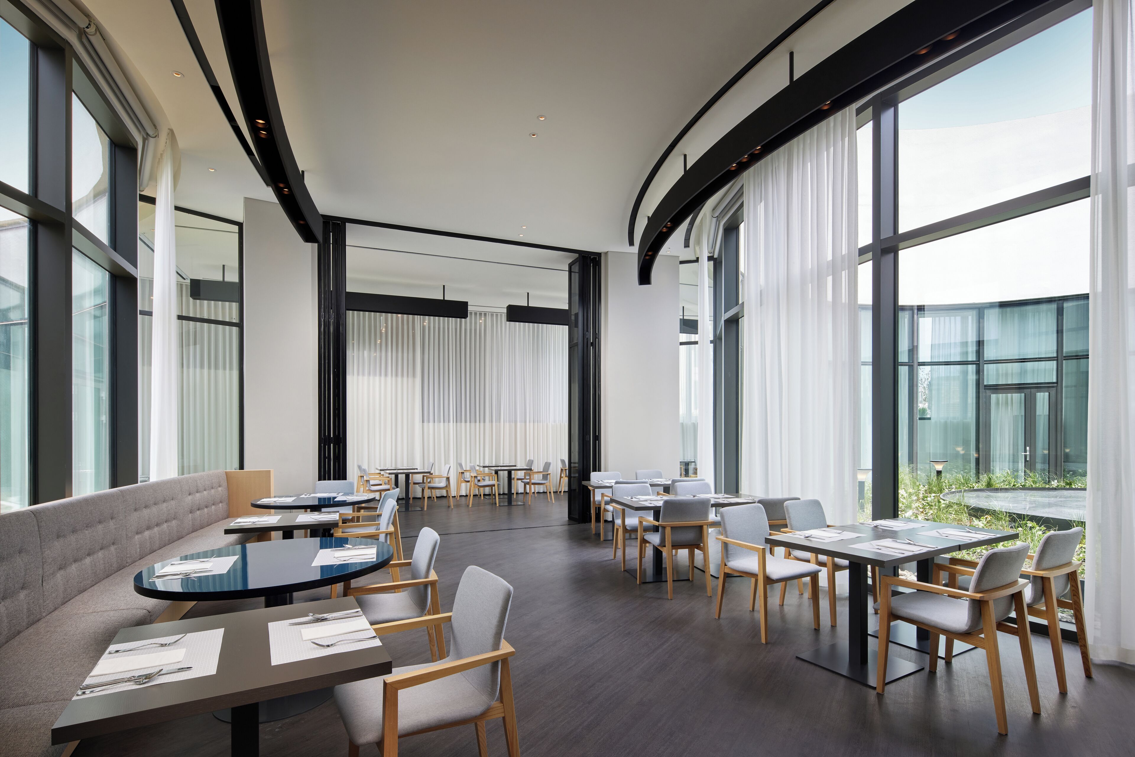 A modern dining area with round tables and grey upholstered seating, large windows, and a view of greenery outside.
