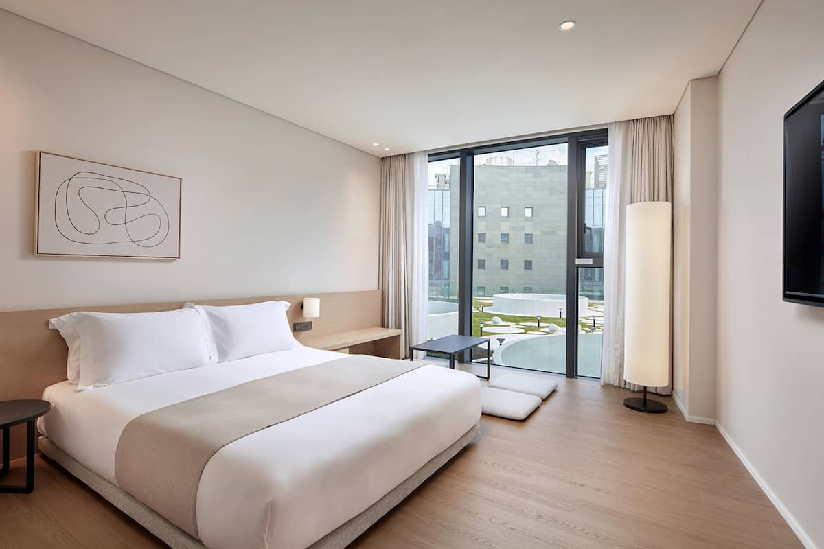A modern hotel room with a large bed, a bedside table, a small table, a lamp, and a view of the city through a large window.