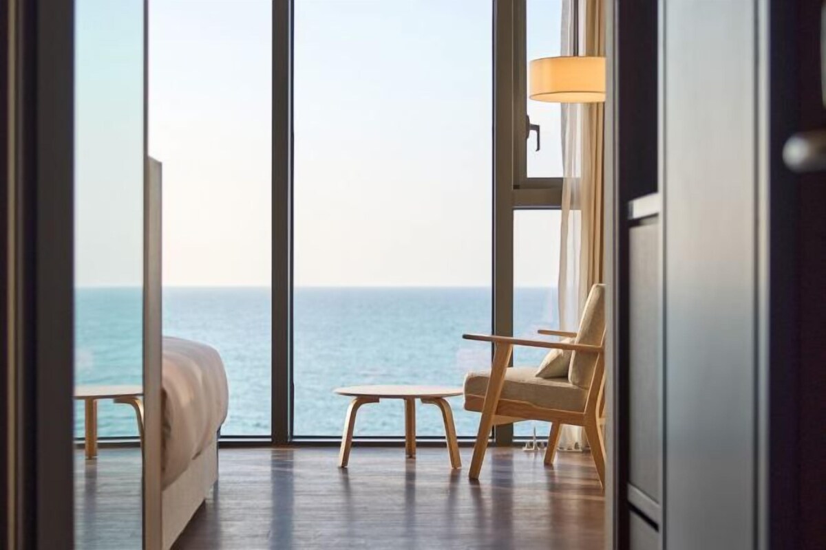 A modern room with a large window overlooking the sea, a sofa, a wooden table, and a chair.