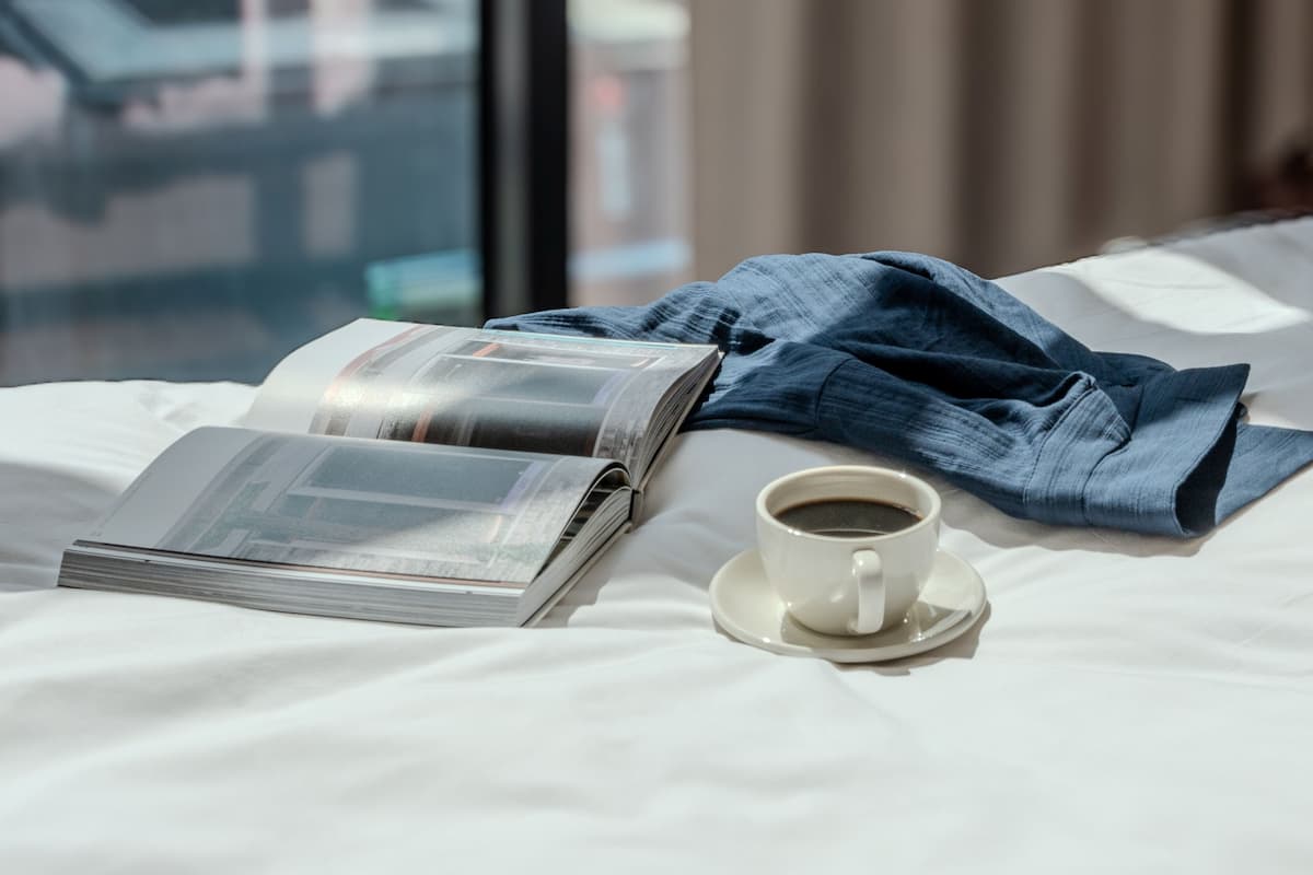 A neatly made bed with a magazine, a cup of coffee, and a pair of jeans on it.