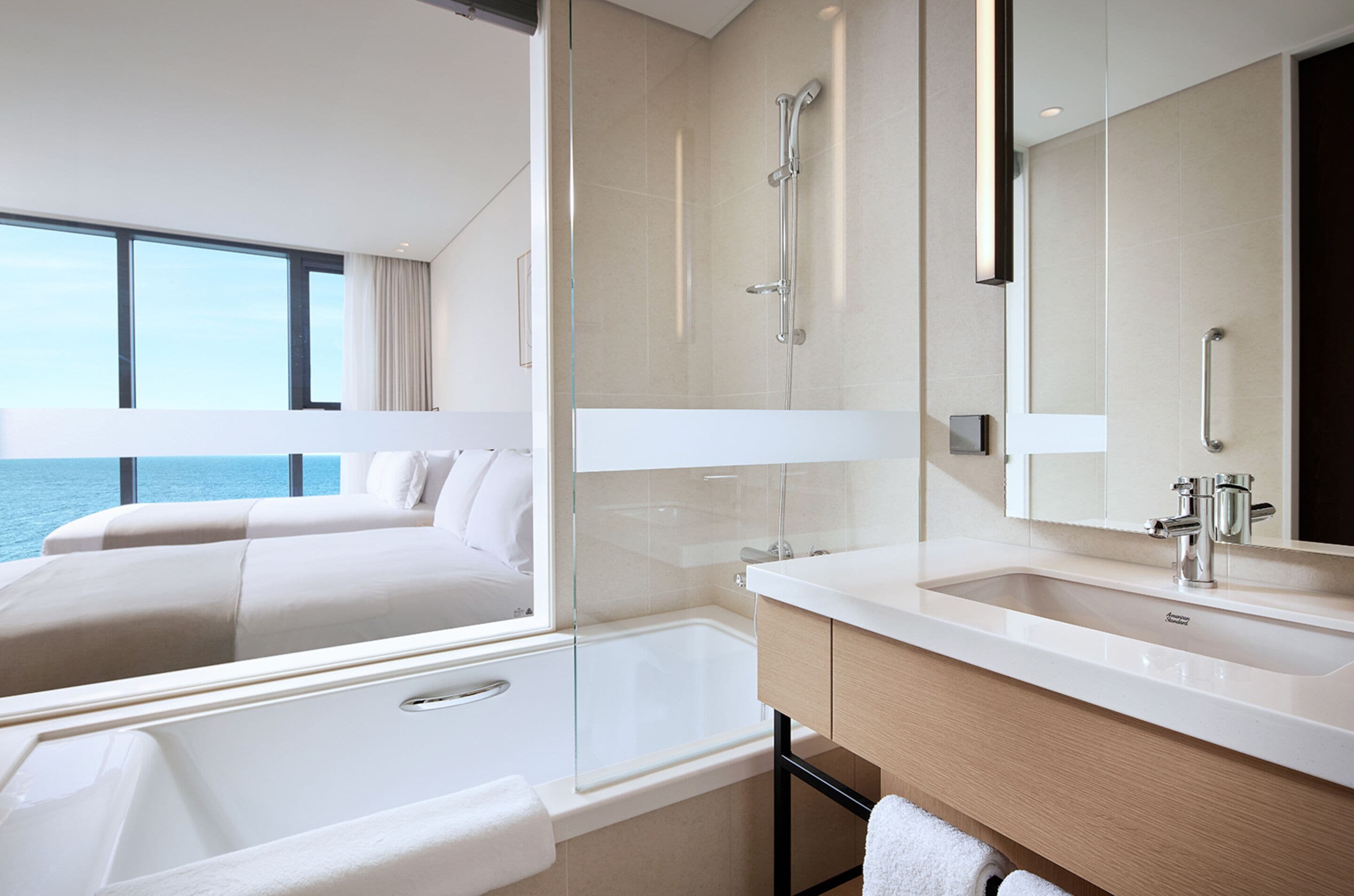 A modern hotel room with a large window, a bathtub, a shower, a vanity with a mirror, and a bed with a view of the sea.