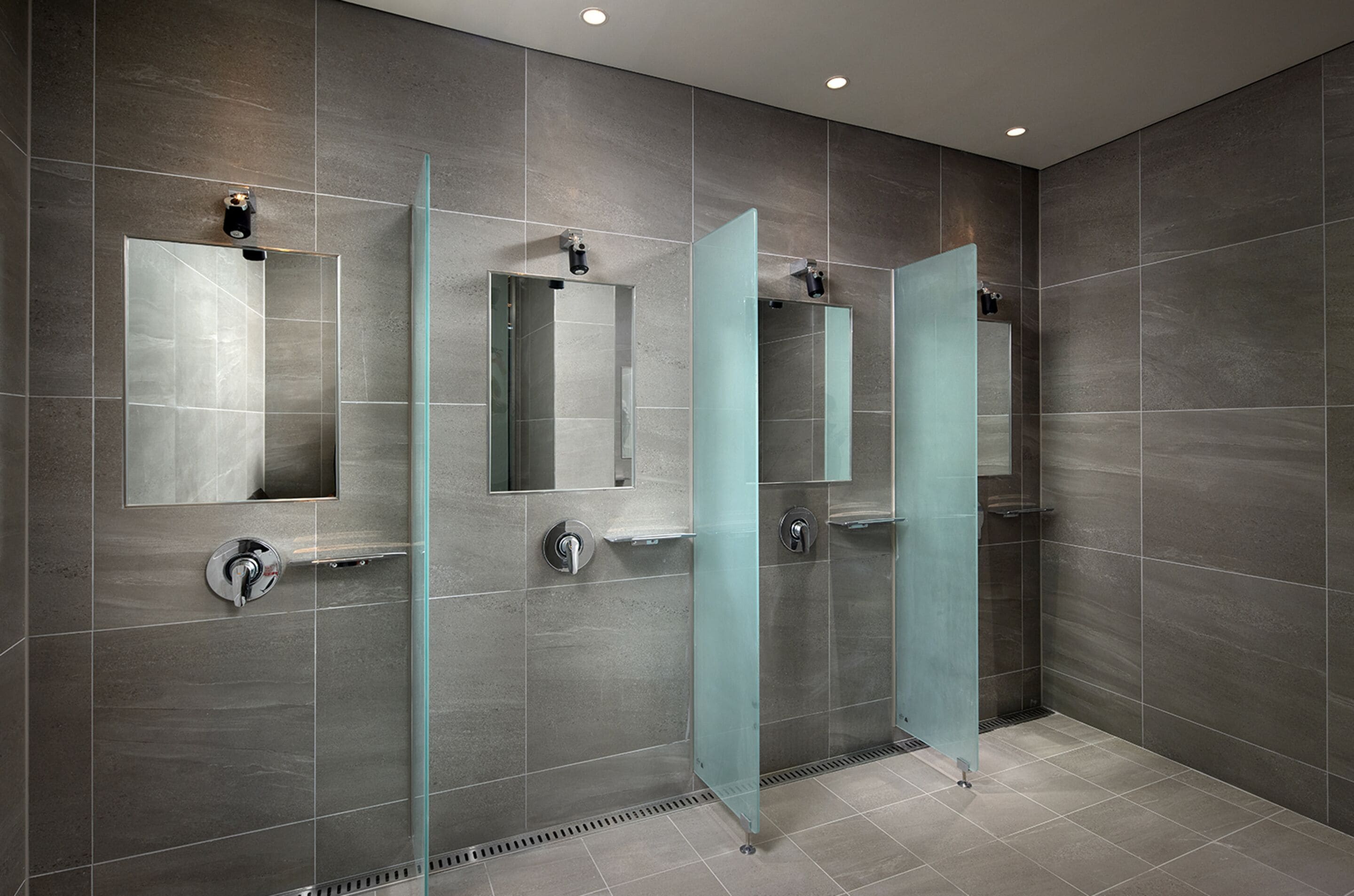 A modern bathroom with three glass-enclosed shower stalls, each with a mirror and a towel rack.