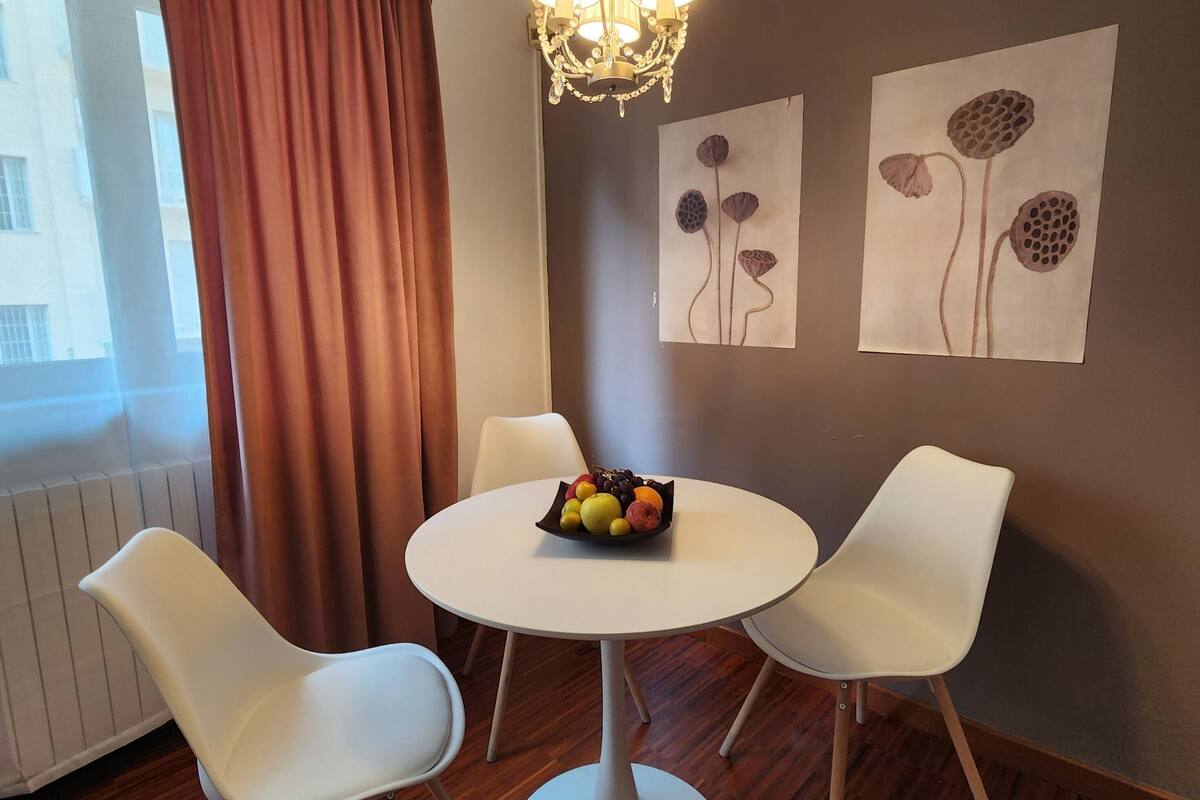 A dining area with a round table, two white chairs, a fruit bowl, a chandelier, and wall art.