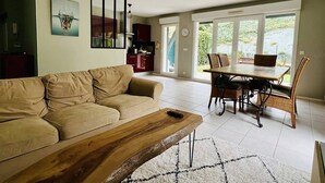 Amplia sala de estar con sofá, mesa de centro, mesa de comedor y sillas. Se observa una cocina al fondo con armarios y un refrigerador.