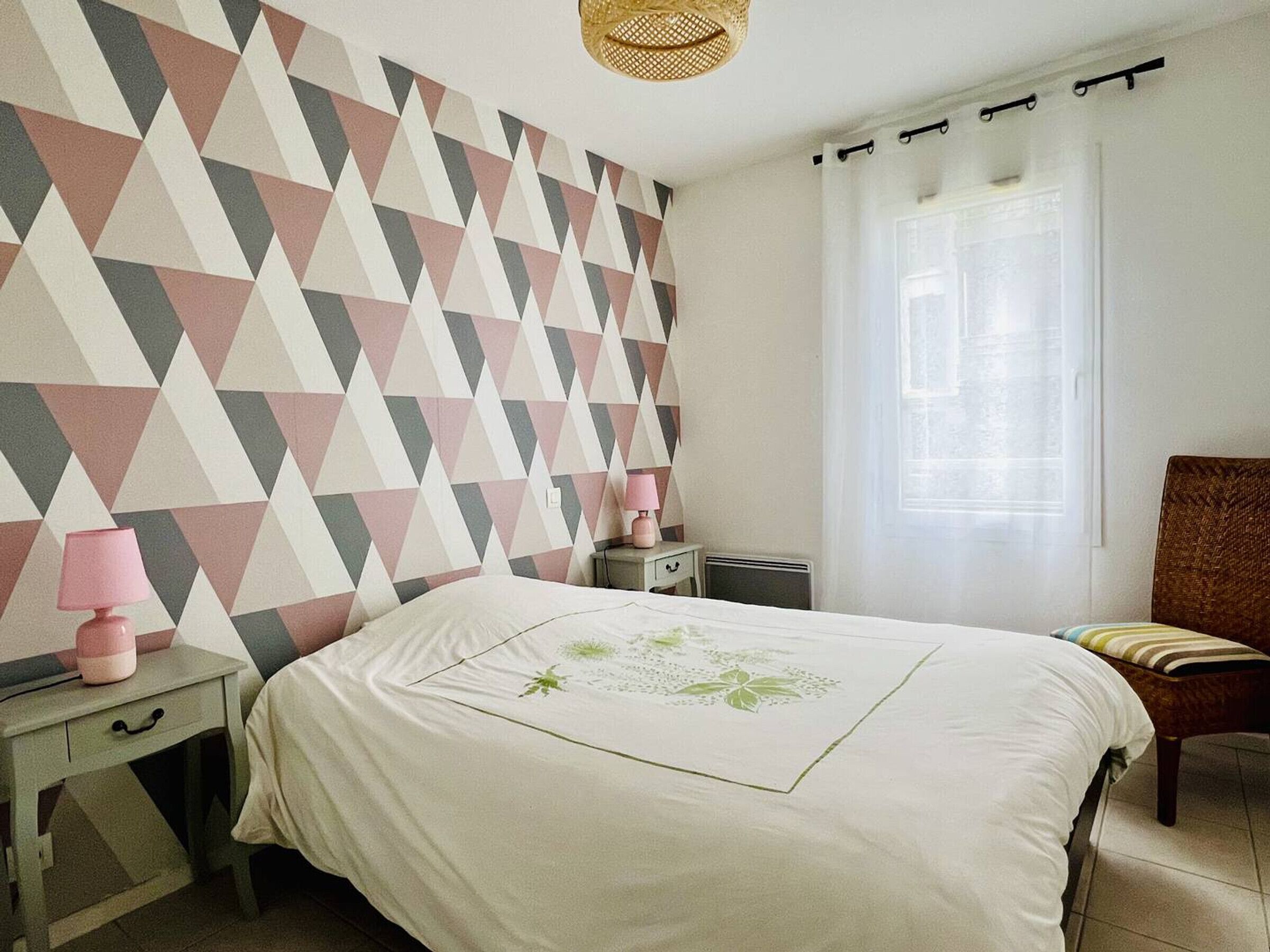 Quarto com parede estampada em padrão geométrico, cama com colcha floral verde, mesa de cabeceira com abajur rosa, uma cadeira e uma janela com cortinas.