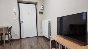 A room with a wooden floor, a white door, a white radiator, a TV on a wooden stand, and a chair. - TWO-ROOM APARTMENT with kitchen-PAVIA-near the center (Pavia)