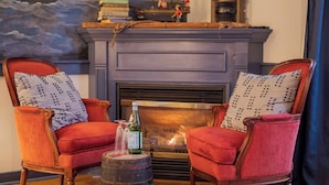 Un salón acogedor con chimenea, dos sillones tapizados en rojo con cojines estampados, una mesita con una botella y vasos, y un cuadro con motivos marítimos sobre la chimenea.
