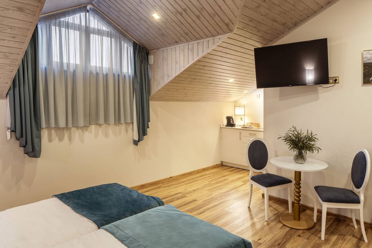 A hotel room with a bed, a desk with a chair, a TV mounted on the wall, and a window with curtains.