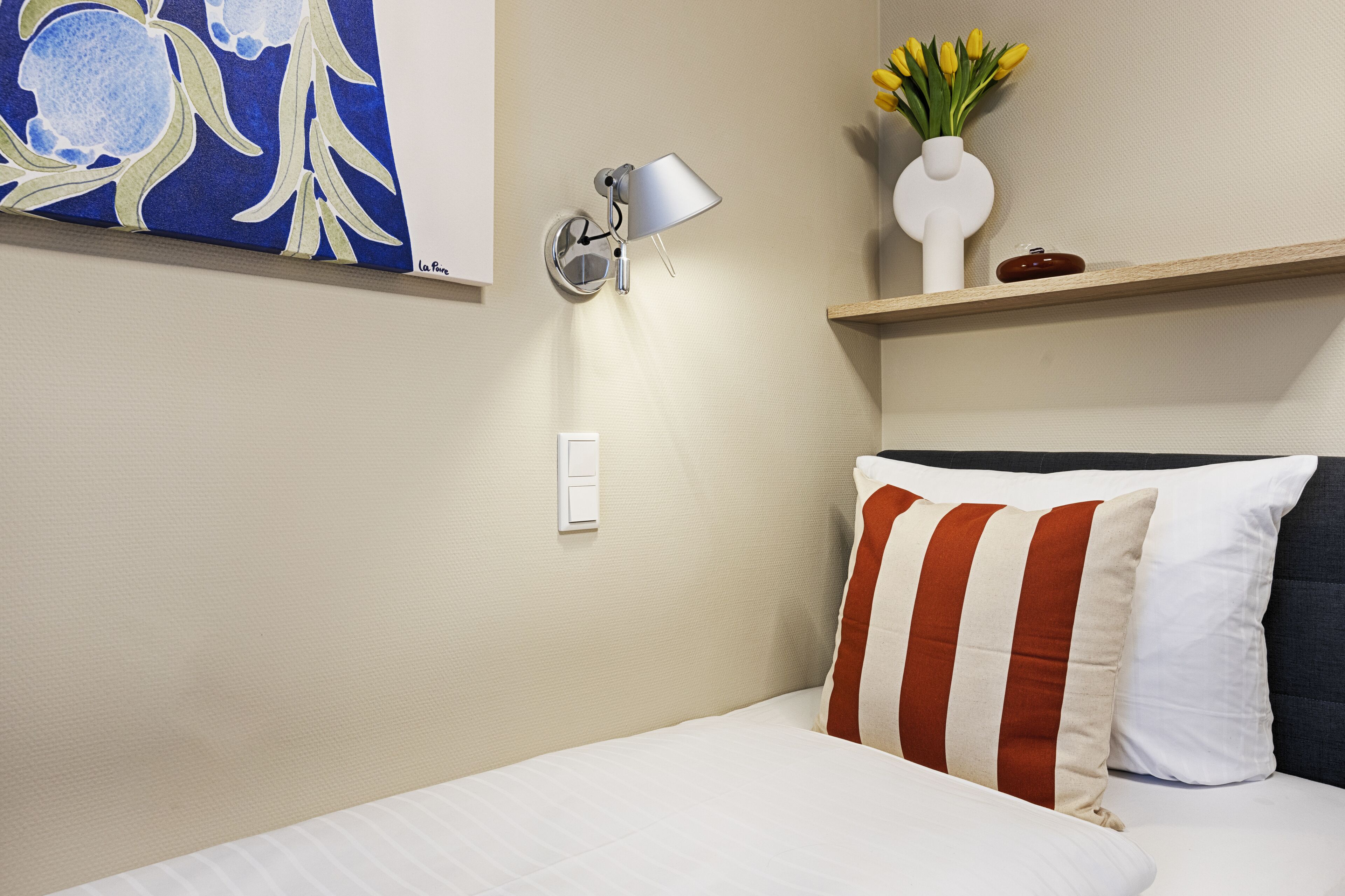 Quarto de hotel com cama, luminária na parede, pintura e prateleira com vaso e flores.