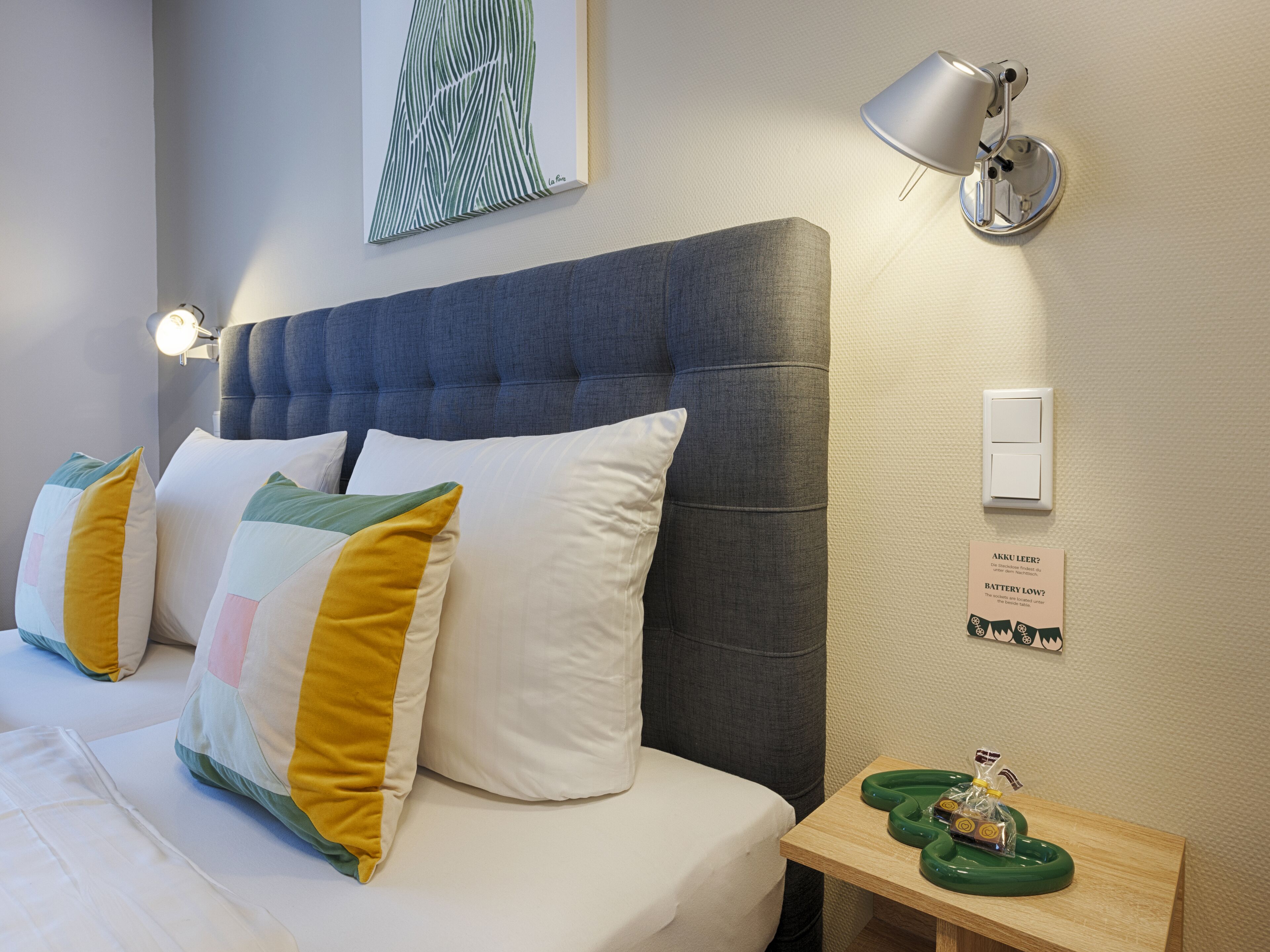 Habitación de hotel con cama, almohadas, mesita de noche, lámpara e interruptor de luz en la pared.