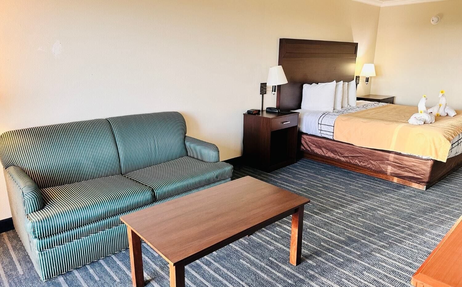 A hotel room with a striped sofa, a wooden table, a bed with a yellow bedspread, a nightstand with a lamp, and a wall-mounted TV.