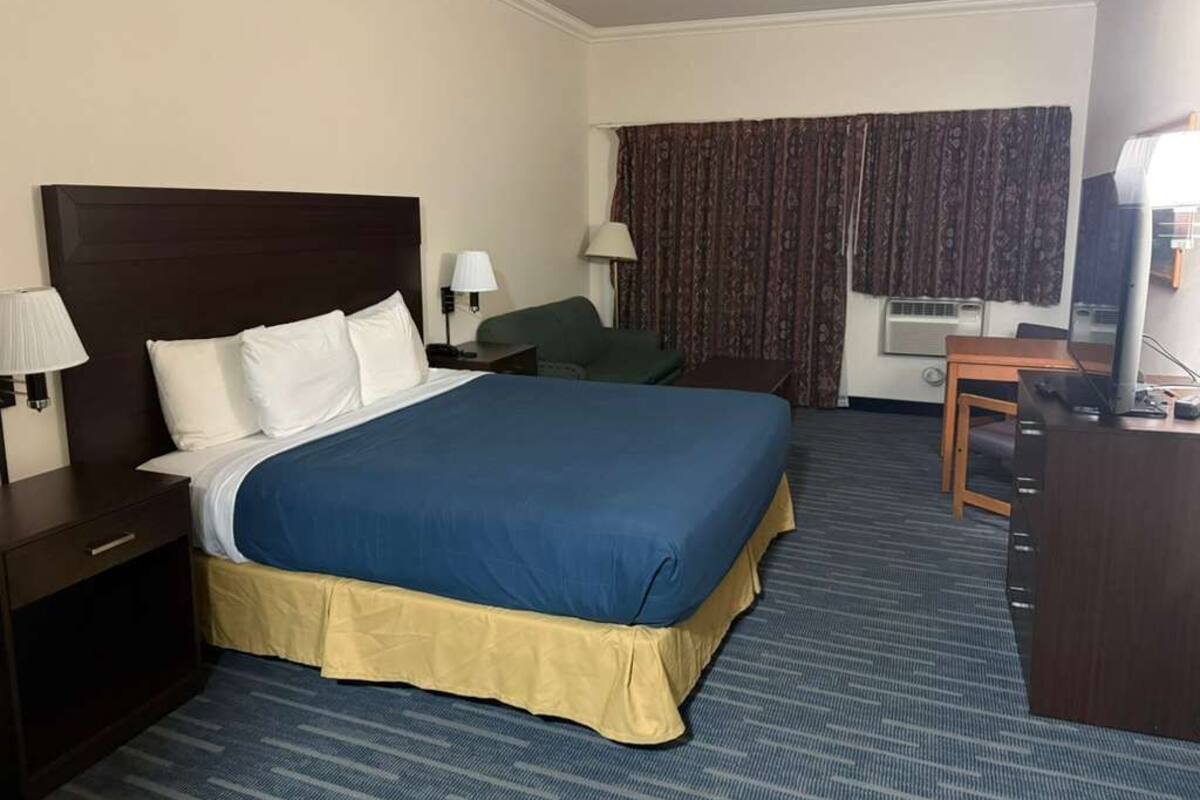 Une chambre d’hôtel avec un grand lit, un bureau avec une chaise, une lampe et une fenêtre avec des rideaux.