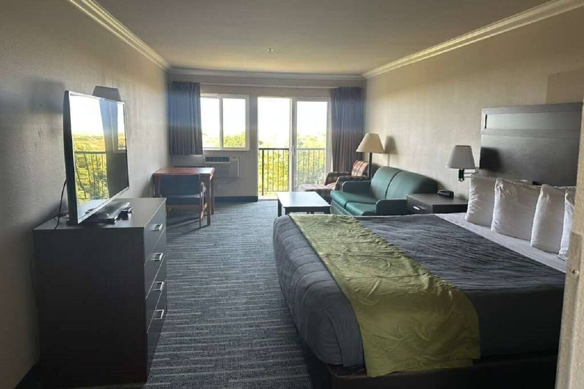 Une chambre d’hôtel comprenant un lit, un bureau, une chaise, une télévision et un balcon avec des rideaux.