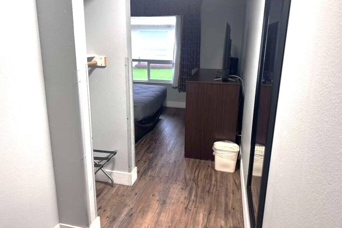 Un couloir étroit mène à une chambre à coucher qui contient un lit, un bureau en bois, une télévision et une fenêtre avec des rideaux.