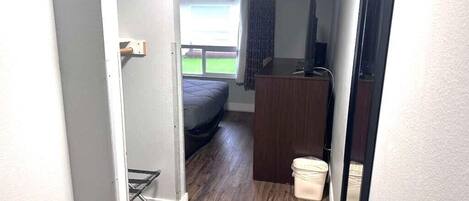 Un pasillo estrecho que da a un dormitorio con cama, un escritorio de madera, televisor y ventana con cortinas.