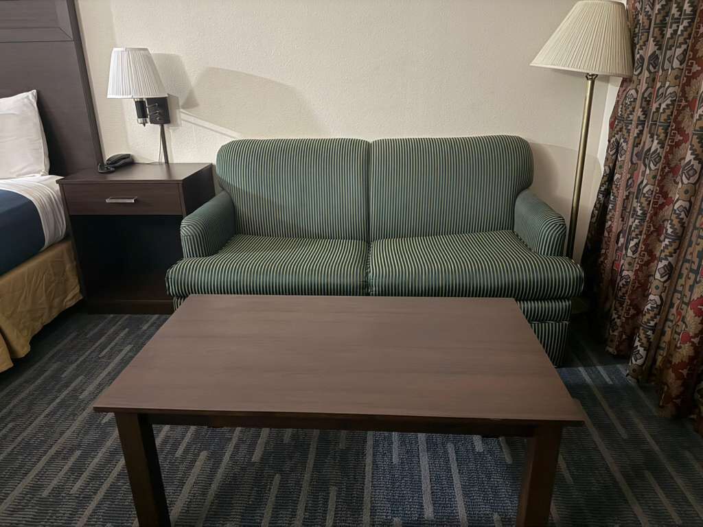A hotel room with a green striped sofa, a wooden coffee table, a patterned carpet, a nightstand with a lamp, and a patterned curtain.
