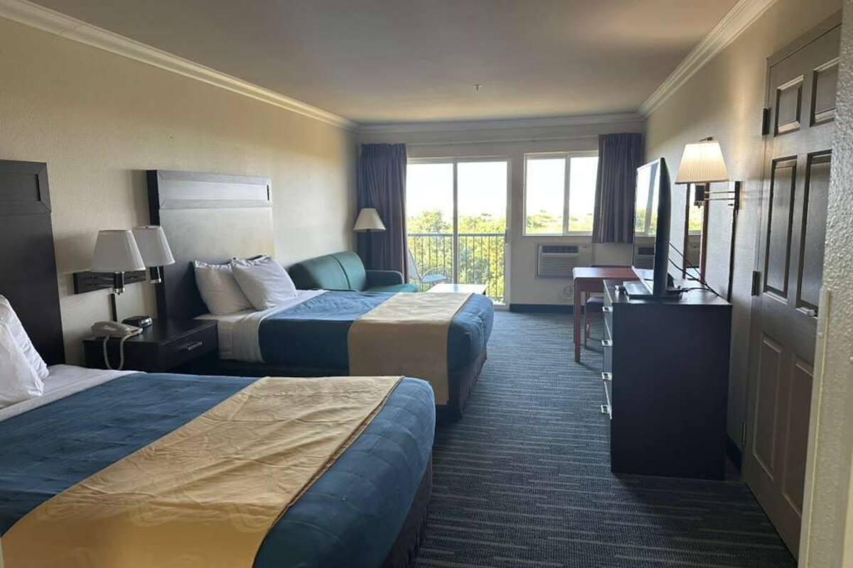 Une chambre d’hôtel avec deux lits, un bureau, une chaise, une télévision et un balcon avec des rideaux.