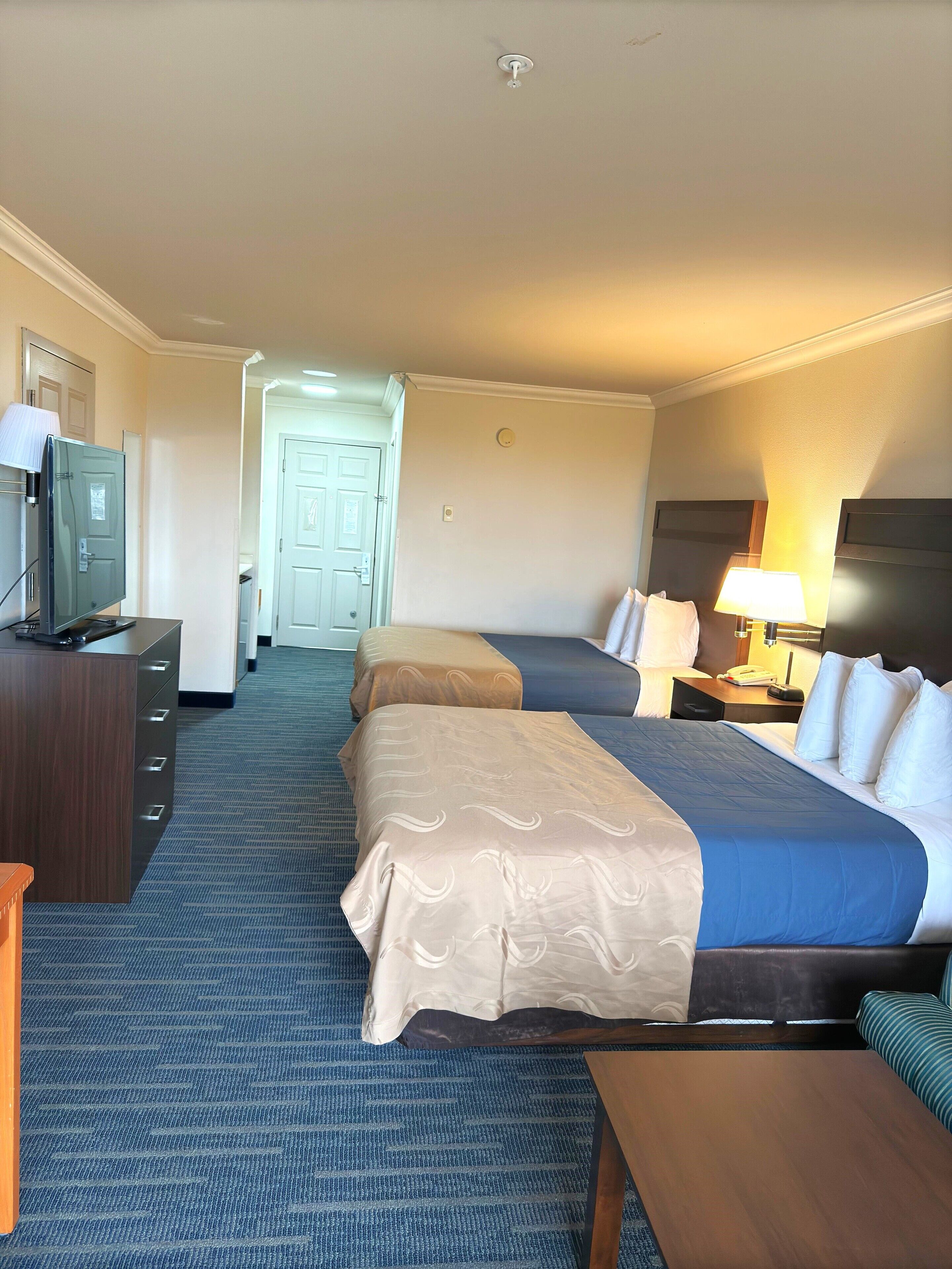A hotel room with two beds, a desk with a TV, a lamp, and a door leading to another room.