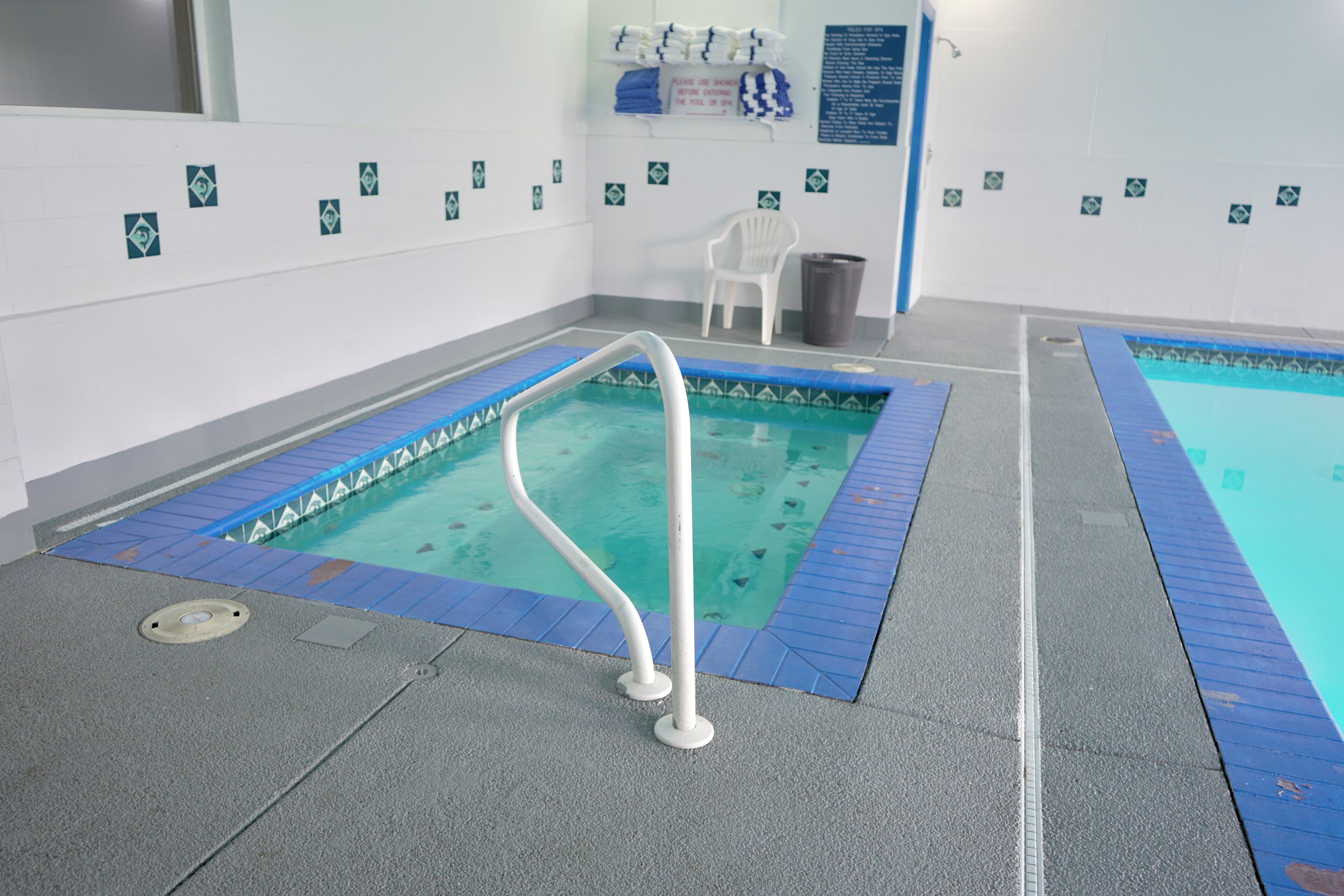 An indoor swimming pool with a blue tiled edge, a white pool ladder, and a white chair in the background.