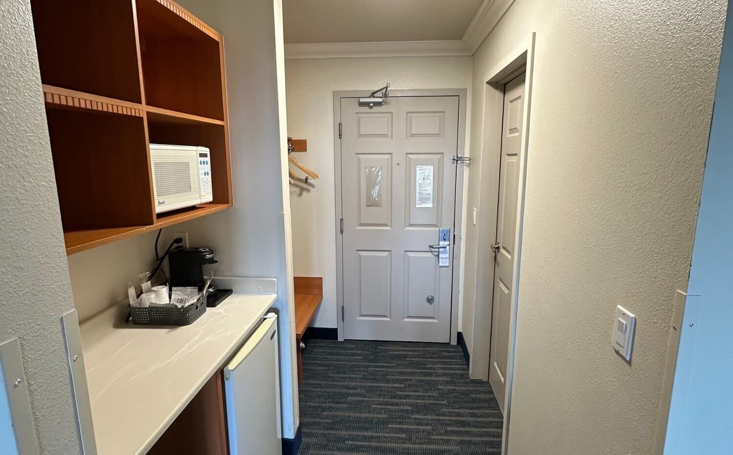 A narrow hallway with a white door at the end, a small desk with a computer and coffee maker, and a shelf with a paper and a small box.