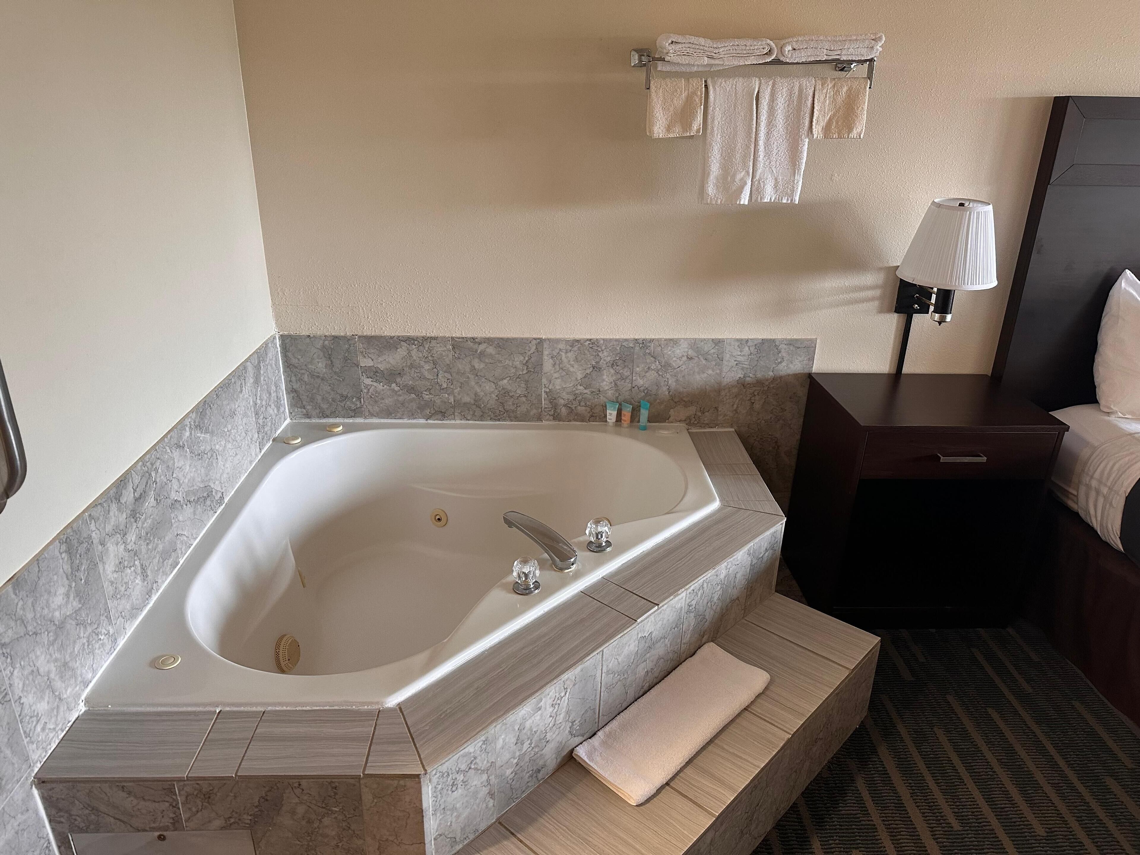 A modern bathroom with a large freestanding bathtub, marble-tiled walls, a wooden bench, a bedside table with a lamp, and a towel rack.