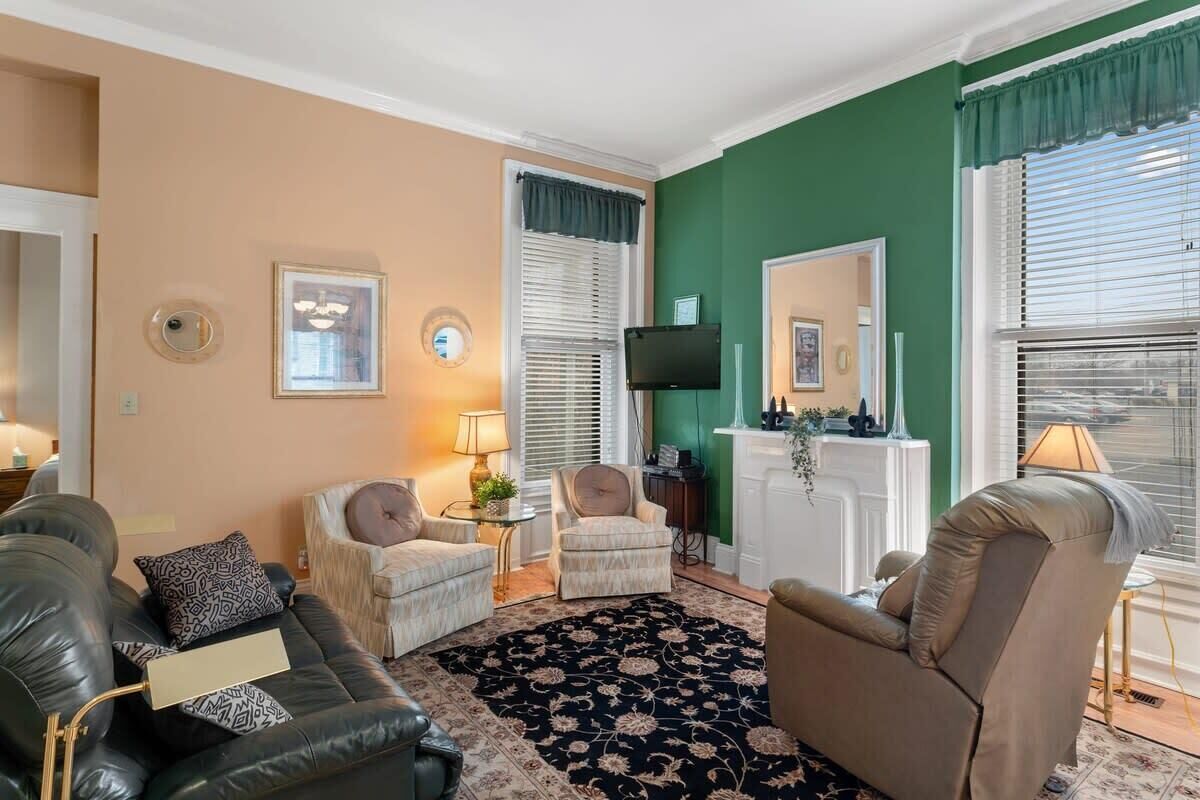 En stue med en beige væg, en grøn accentvæg og en sort blomsterløber. Der er to lænestole, en sofa og et fjernsyn ophængt på væggen.