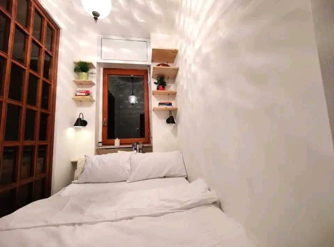 A narrow room with a bed, wooden shelves, and a window with a view.
