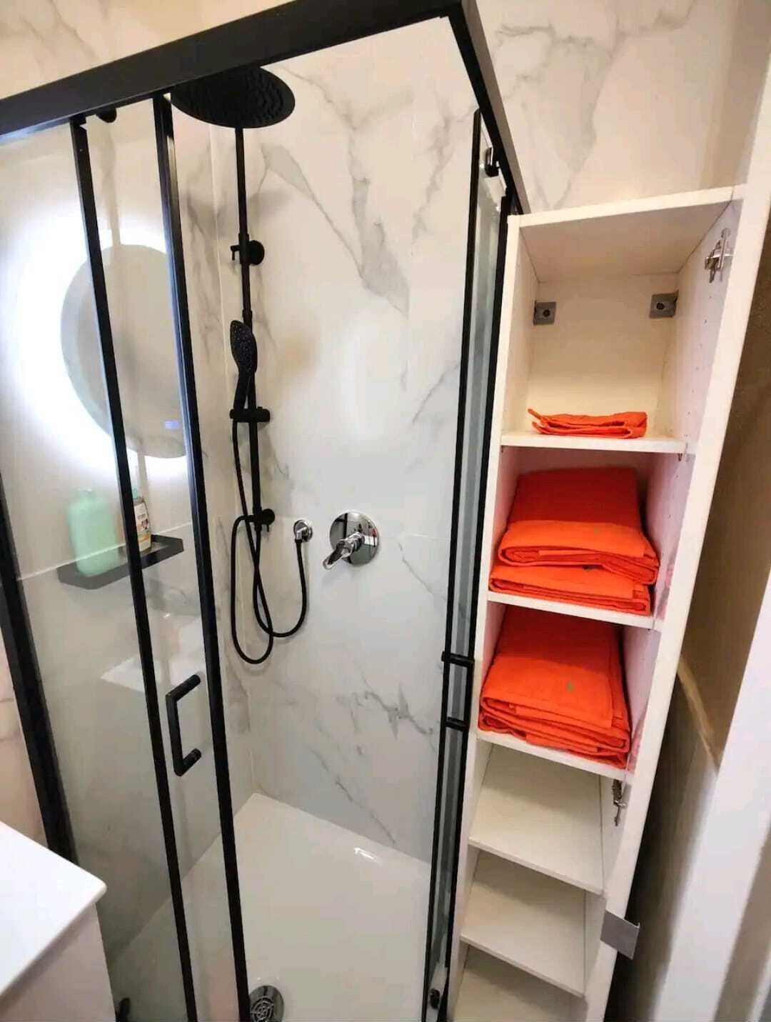 A modern bathroom with a glass-enclosed shower, marble walls, and a white shelf with folded towels.