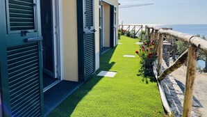 A row of hotel rooms with open shutters, a grassy pathway, and a wooden fence overlooking the sea. - Eufonia del Mare (Finale Ligure)