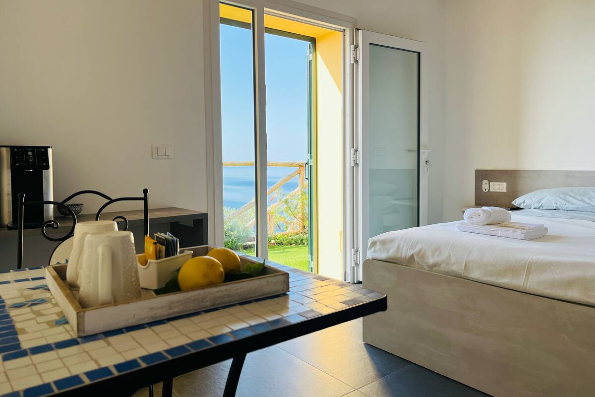 A hotel room with a bed, a table with a tray of lemons, a coffee machine, and a view of the sea through a sliding glass door.