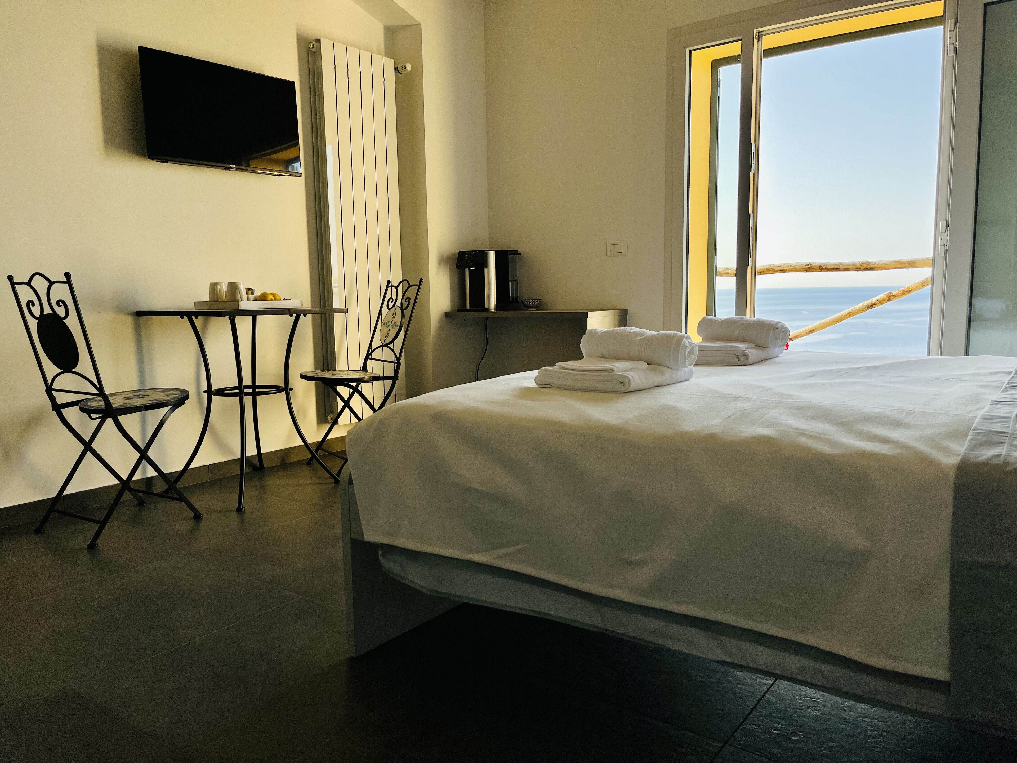 A hotel room with a bed, two chairs, a small table, a TV, and a window with a view of the sea.