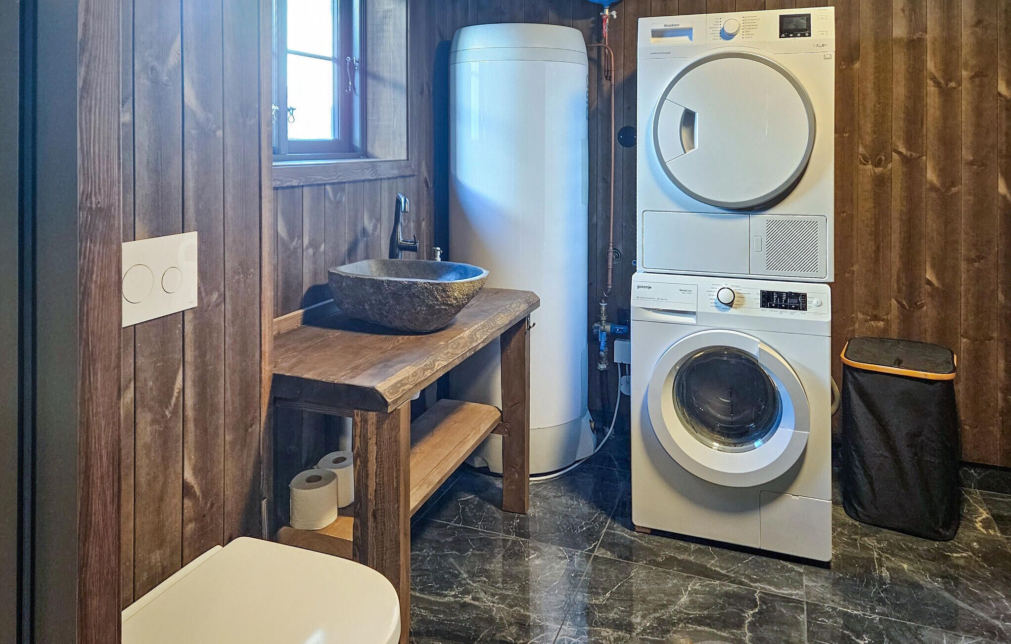 A compact laundry room with a washing machine, a large water heater, a wooden shelf with a basin, and a toilet.