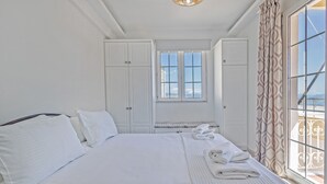Une chambre d’hôtel avec un grand lit, du linge de lit blanc, une fenêtre donnant sur un beau paysage et un balcon agrémenté de rideaux à motifs.