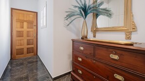 Un couloir avec une porte en bois, un sol en marbre, une commode en bois dotée de tiroirs, un vase orné de plumes bleues et un miroir à cadre doré.