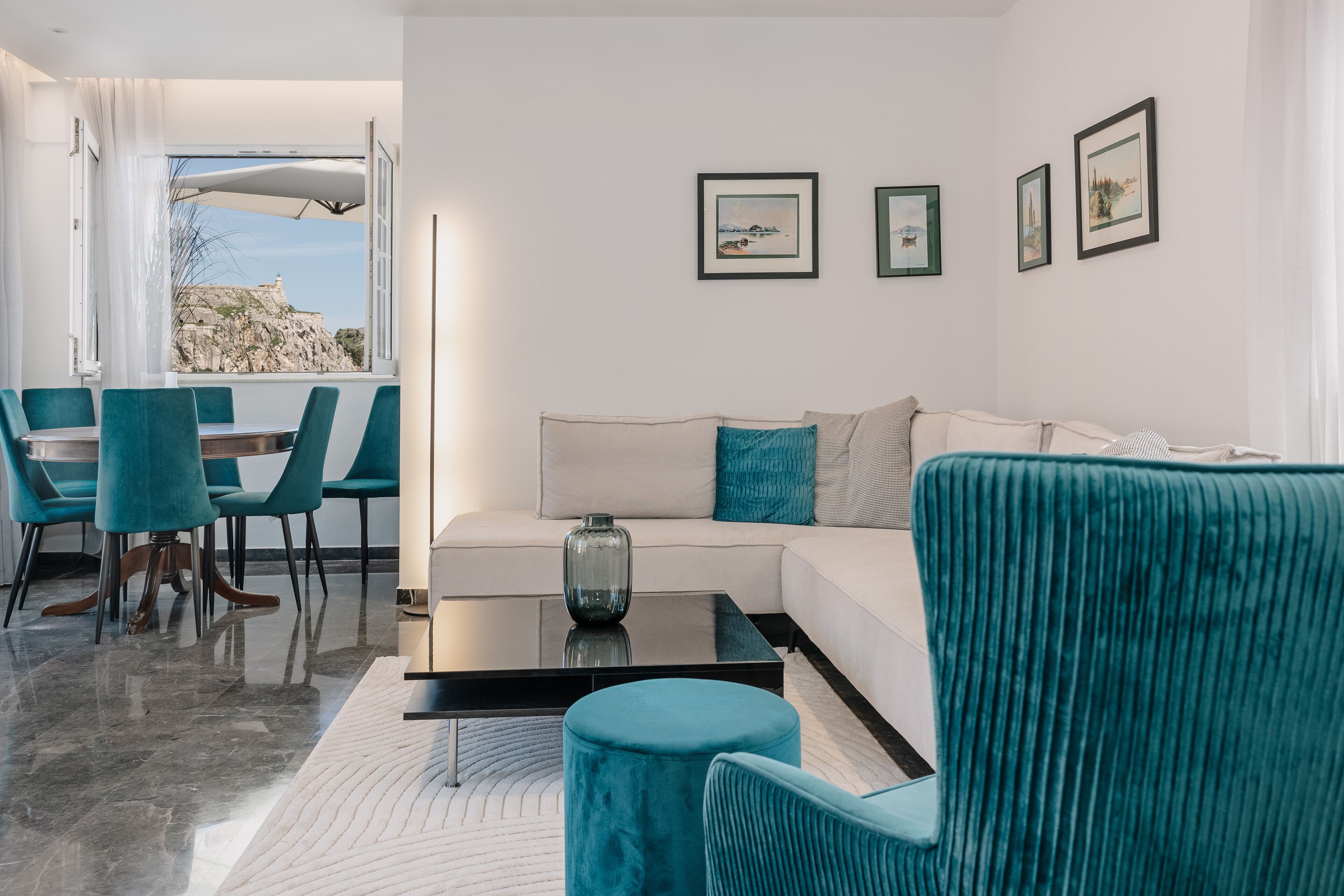 Uma sala de estar moderna com um sofá branco, cadeiras verde-água e uma mesa de centro de vidro. Há quadros nas paredes e uma vista de uma paisagem rochosa pela janela.