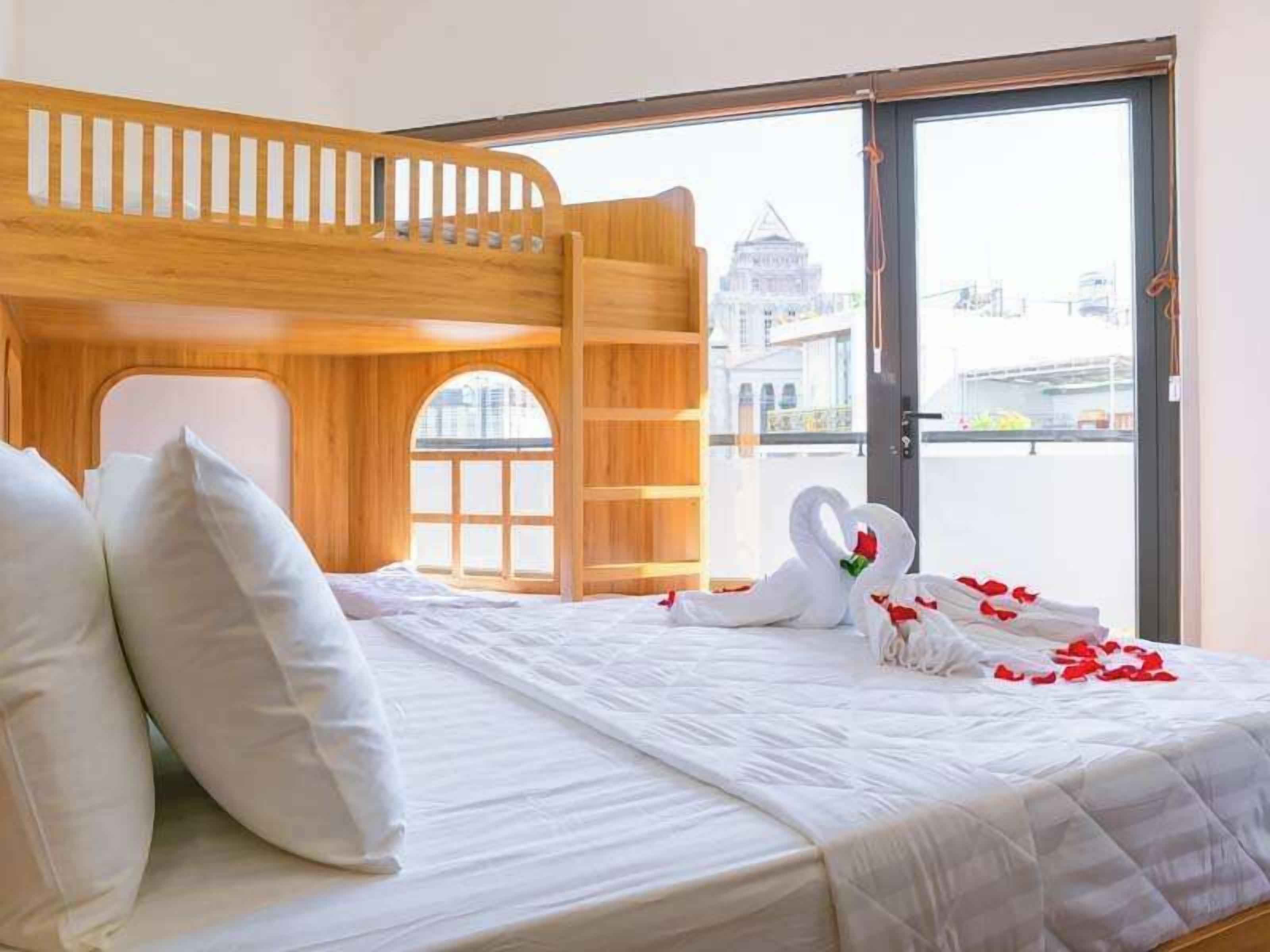 A hotel room with a bunk bed, a large window with a view of a cityscape, and a bed adorned with white bedding and rose petals.