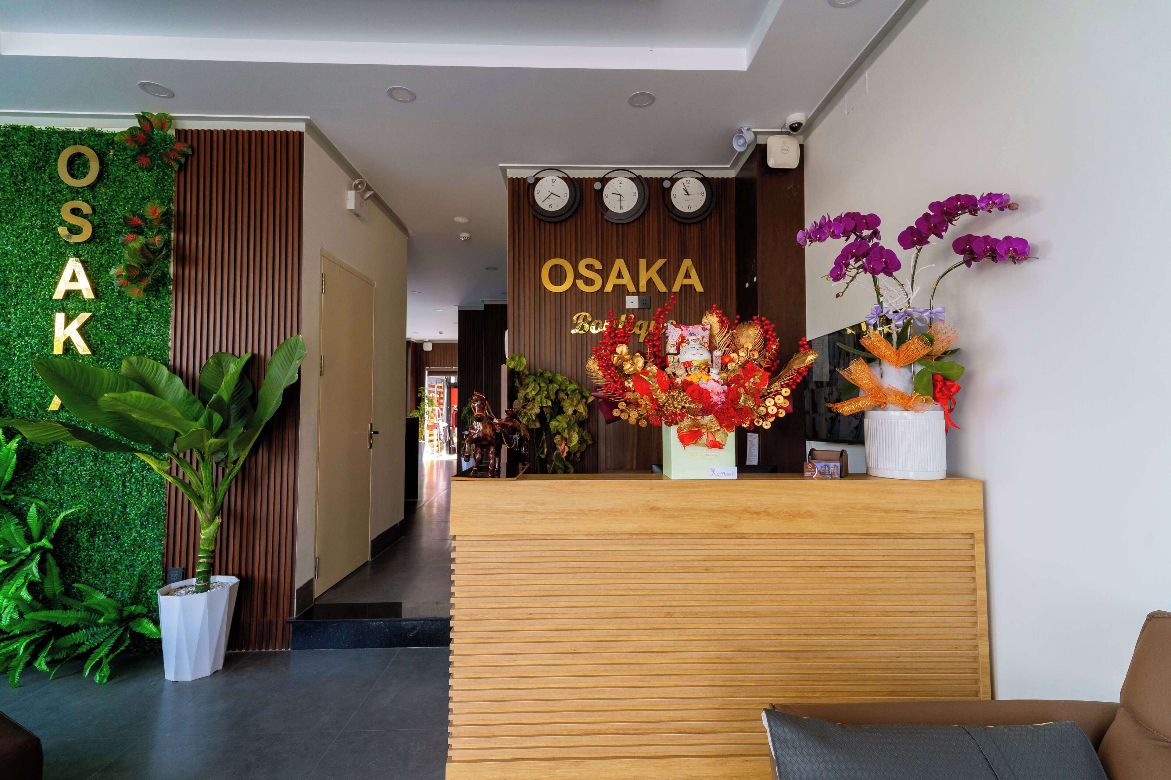 A reception area with a wooden desk, a green wall with 'OSAKA' signage, and decorative flowers.
