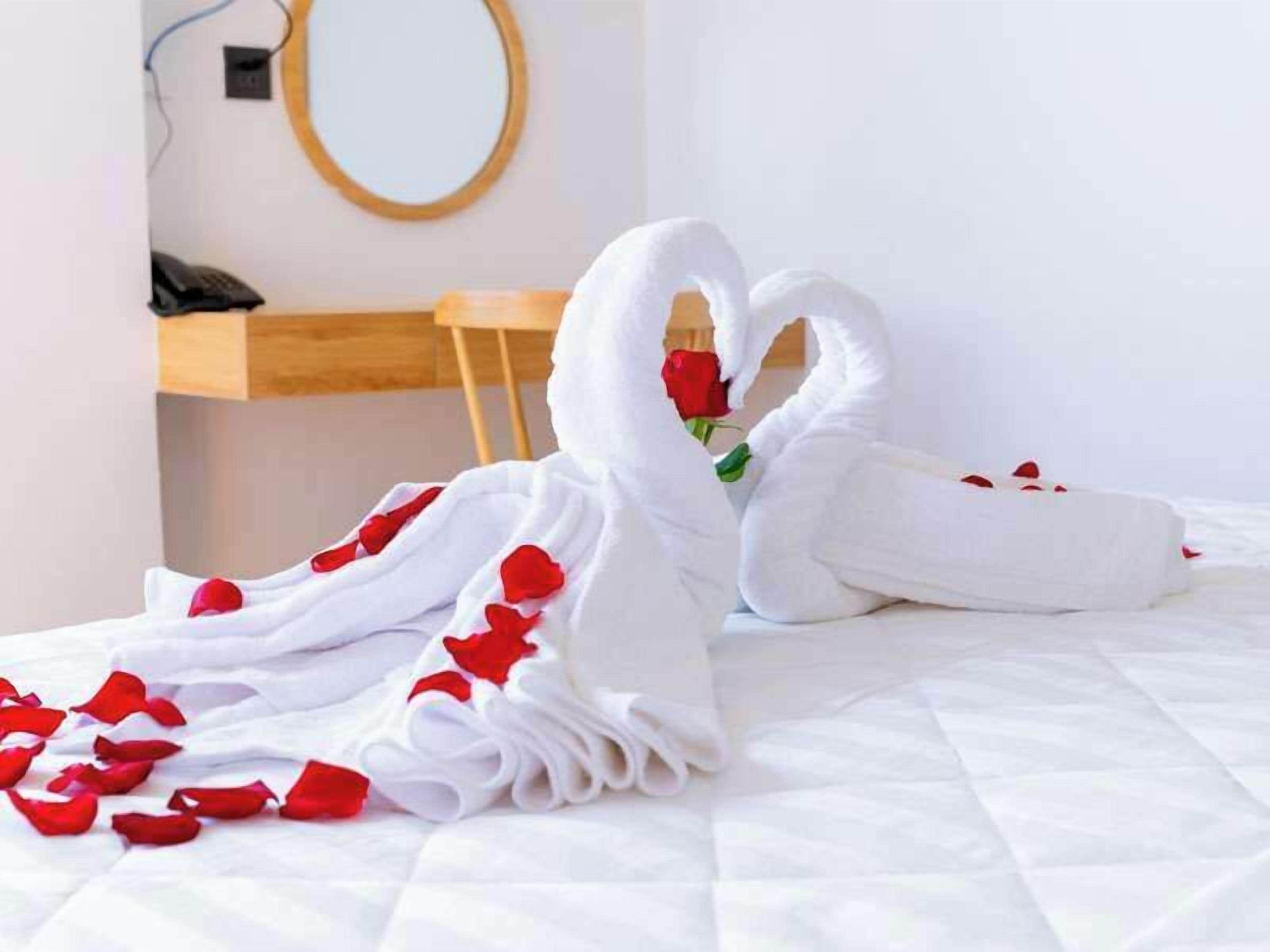 Una cama con sábanas blancas y dos toallas con forma de cisne adornadas con pétalos de rosa roja.