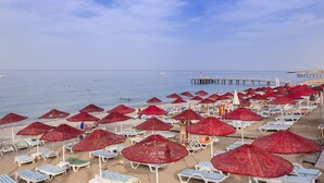 A beach with rows of sun loungers and red umbrellas, a pier in the background, and calm sea. - Holiday Garden Resort - All inclusive (Alanya)